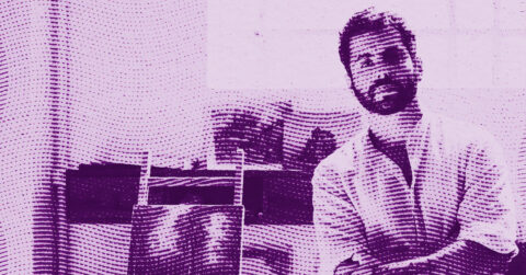 A bearded man sitting and smiling in a room with shelves and books behind him, shown in a purple halftone pattern style. © Illustration by ArtCritic.