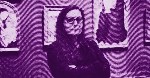 A person with long hair and glasses stands with crossed arms in front of framed artwork on a wall. The image has a purple color filter and a dotted texture effect. © Illustration by ArtCritic.