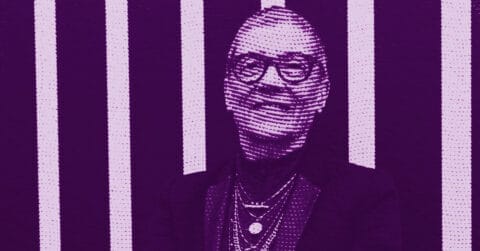 A stylized, purple-tinted image of a person with short hair and glasses, smiling, wearing layered necklaces and a blazer, with vertical white stripes in the background. © Illustration by ArtCritic.
