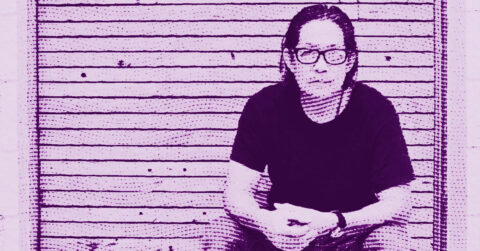 A person with long hair and glasses sits in front of a horizontal wooden slat wall. The image has a purple tint and a halftone effect, giving it a stylized, artistic appearance. © Illustration by ArtCritic.