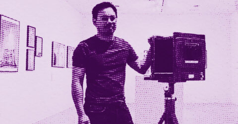 A person stands in an art gallery, smiling and holding a large vintage camera on a tripod. The image has a purple halftone effect, with framed artworks visible on the walls in the background. © Illustration by ArtCritic.