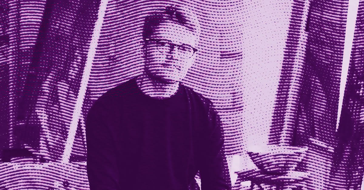 Alfie Caine wearing glasses sits indoors in front of curtains. The image has a purple tint with a wavy, textured filter effect applied, creating a stylized and abstract appearance. © Illustration by Art Critic.