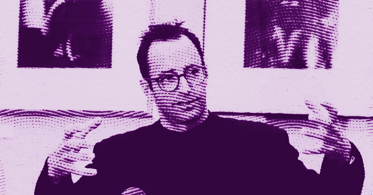 Herb Ritts wearing glasses gestures with his hands while sitting in front of two framed pictures. The image has a purple and dotted graphic effect. © Illustration by Art Critic.